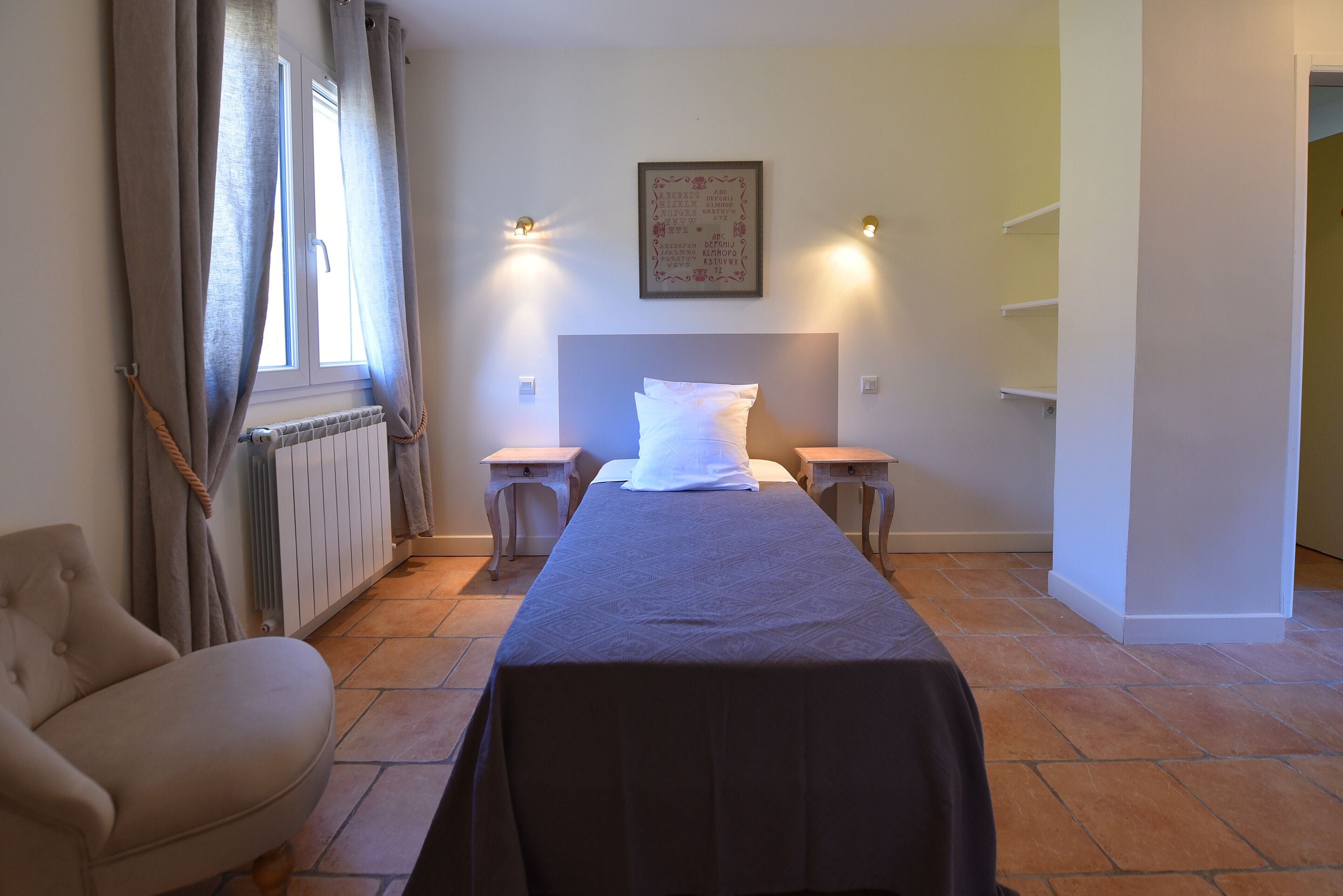 Une chambre d’hôtel avec un lit, une chaise, un radiateur, une fenêtre avec des rideaux et des lampes fixées au mur.