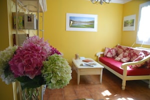 Una habitación con paredes amarillas, suelo de madera, un sofá blanco con cojines rojos, una mesita y un jarrón de flores.
