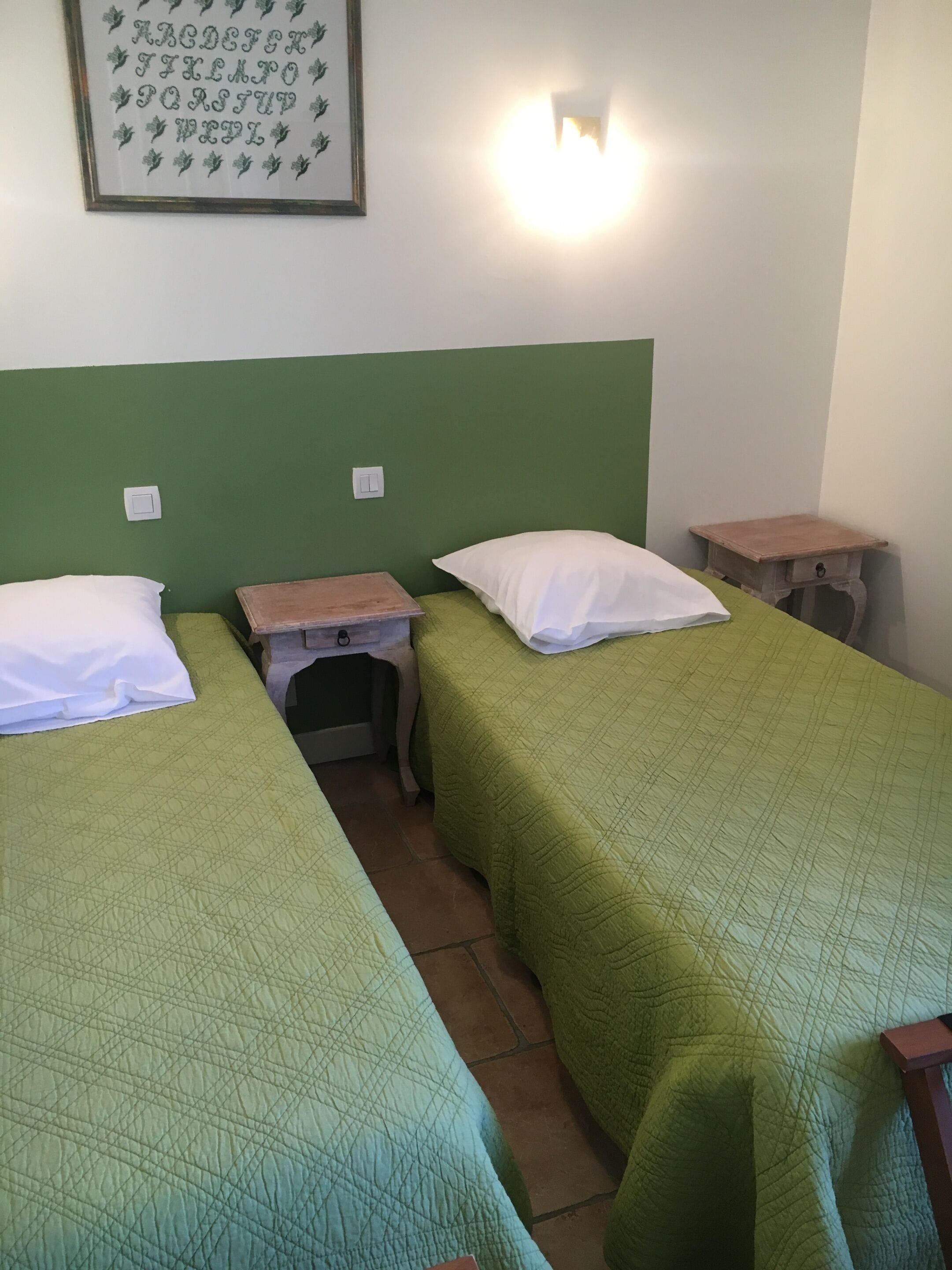 A room with two single beds, each with a green quilted bedspread and white pillows. There is a small wooden nightstand with a lamp between the beds. A framed alphabet poster hangs on the wall above the beds.