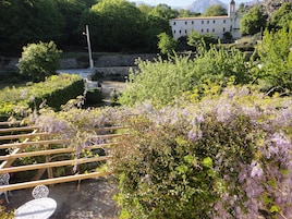 Un jardín con una pérgola y una mesa, rodeado de exuberante follaje y flores moradas.