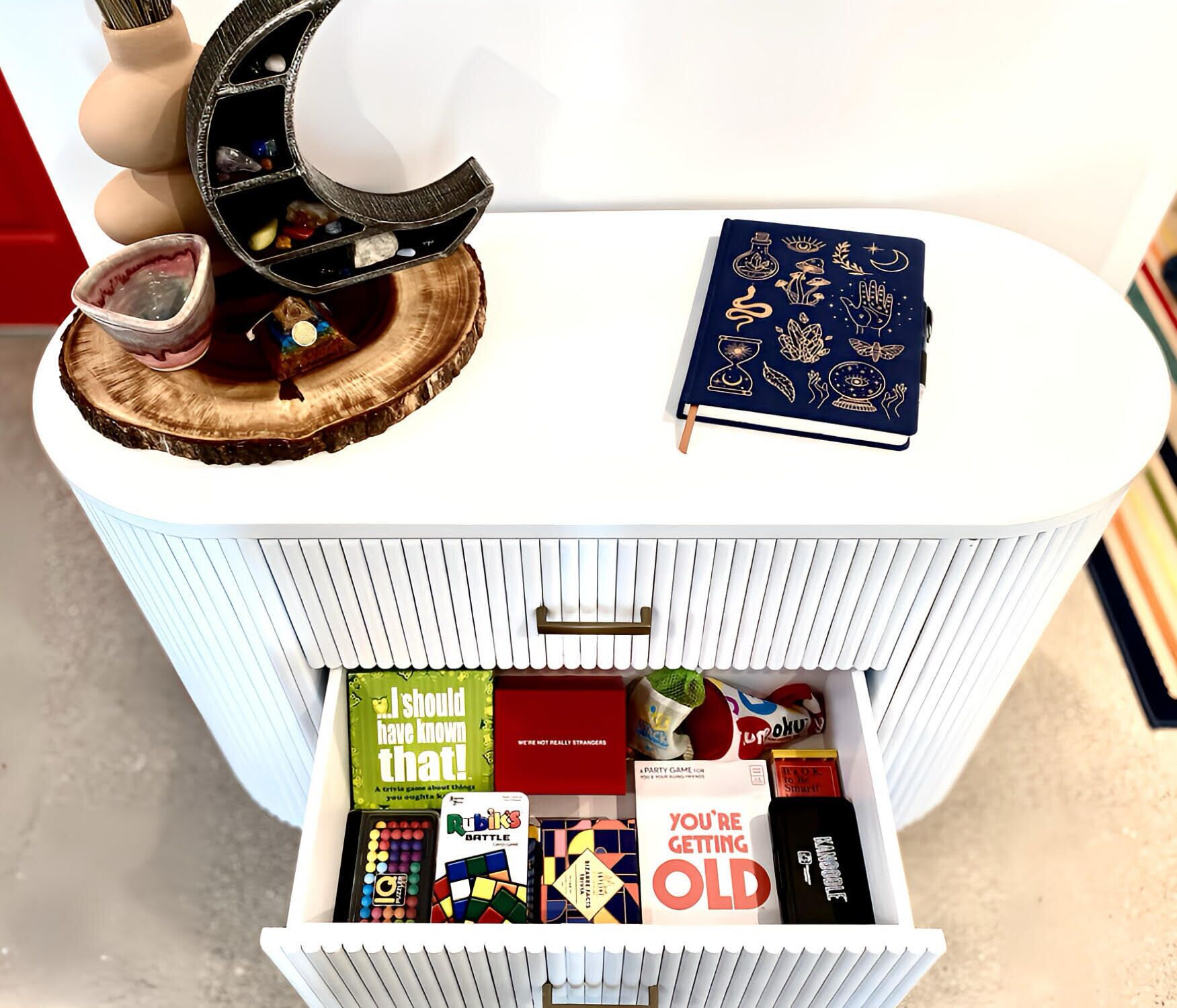 A white storage unit with a top shelf holding a decorative item and a book, and a lower shelf containing various board games.