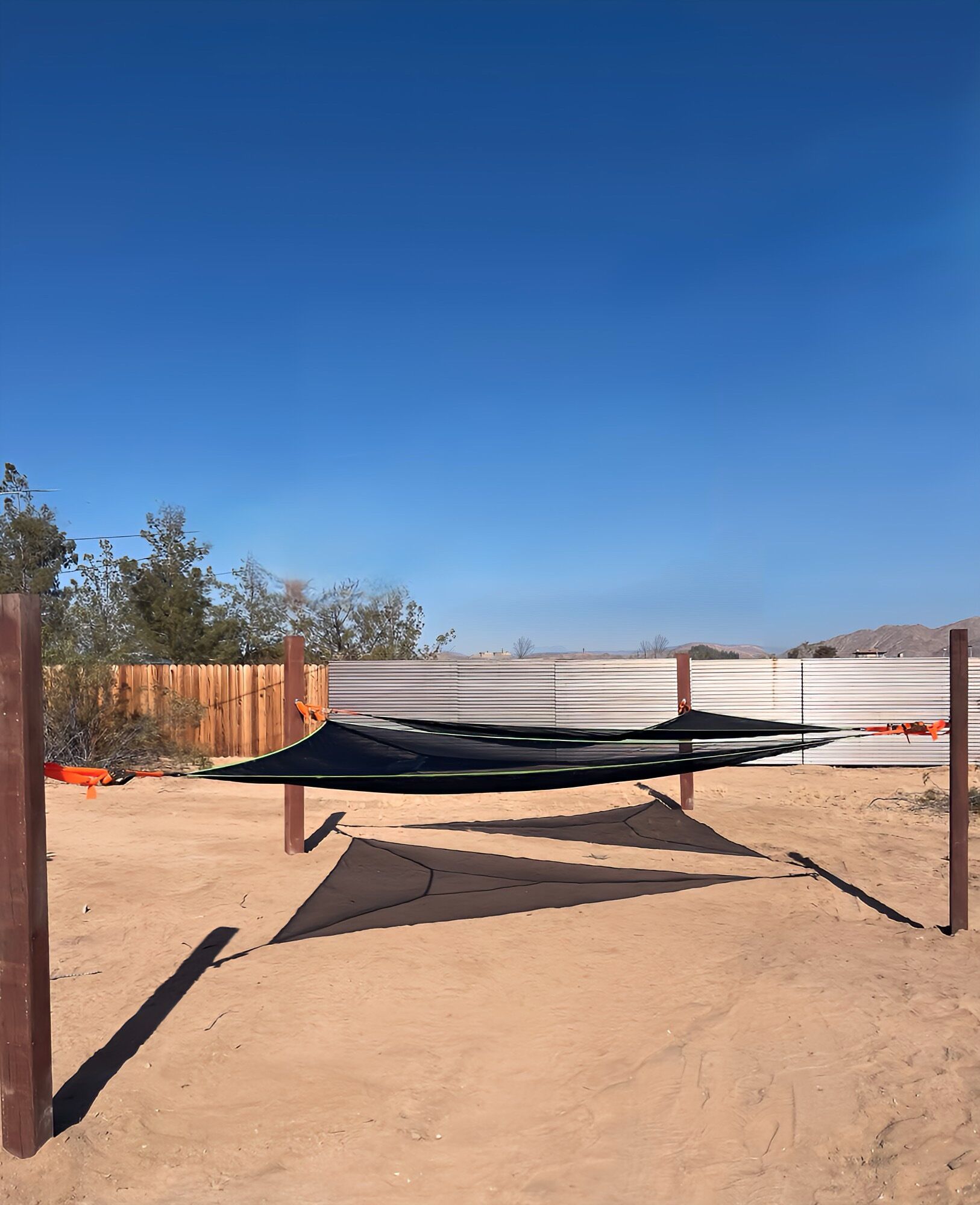 Un hamac est installé sur une surface sablonneuse, avec une clôture en bois et une clôture en métal en arrière-plan.