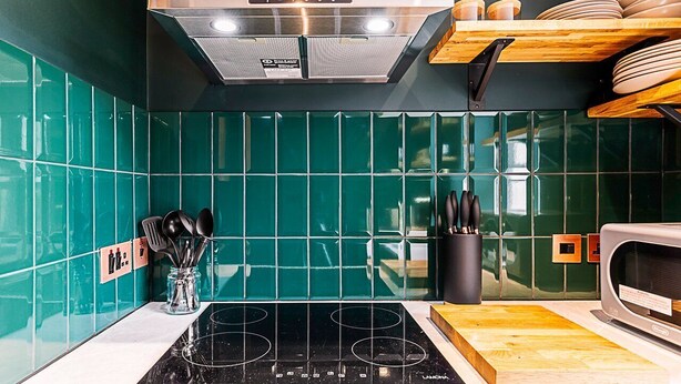 A kitchen with teal tiles, a stainless steel range hood, a microwave, and wooden shelves.