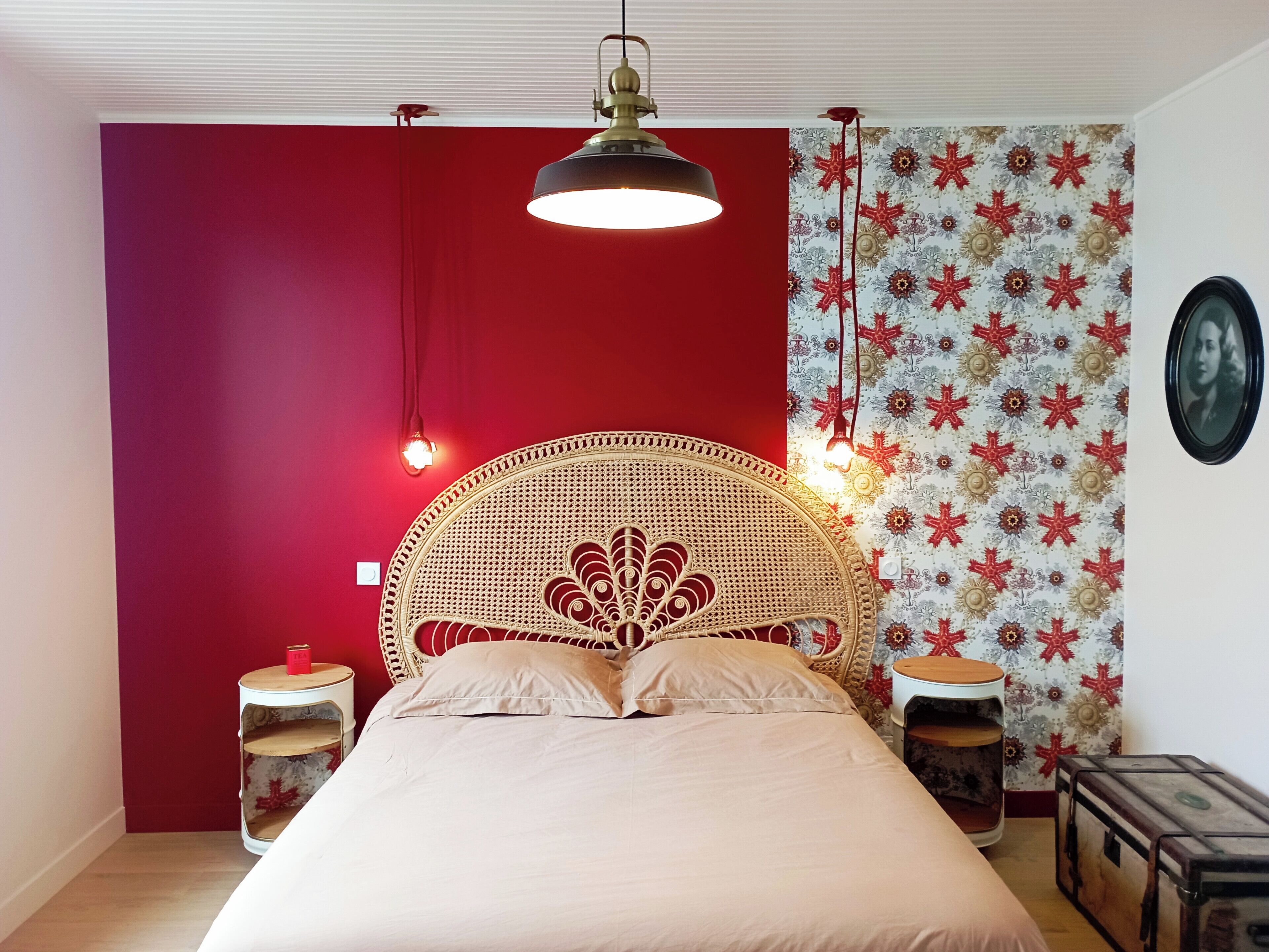 Een slaapkamer met een rode accentmuur, een wit rieten hoofdbord, een bed met beige beddengoed, behang met een patroon, een ronde tafel en een vintage koffer.