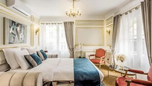 A hotel room with a large bed, a red armchair, a small table with a vase of flowers, and a chandelier.