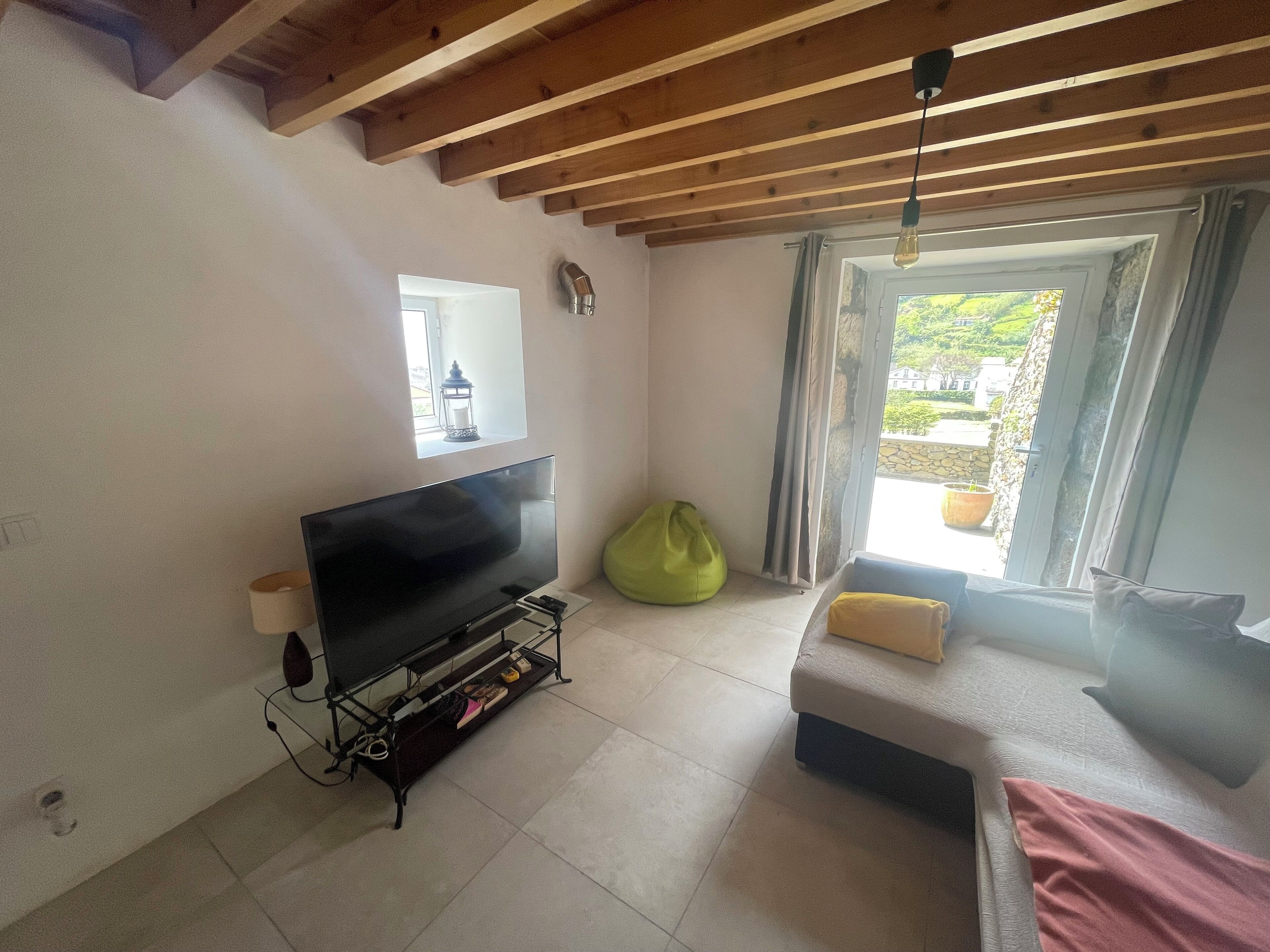 Een kamer met een tv op een standaard, een relaxfauteuil, een bed met kussens, een raam met gordijnen en uitzicht op groen.