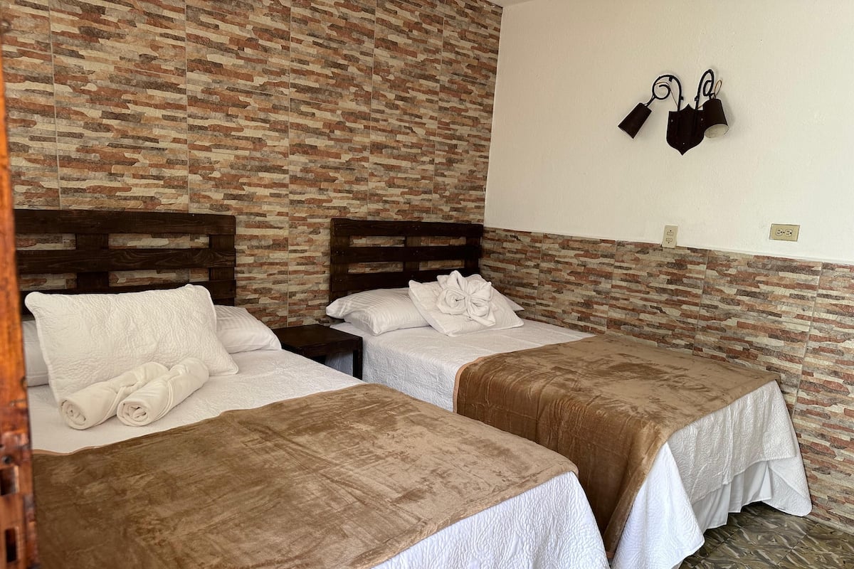 A hotel room with two beds, each with a brown bedspread and white linens. There is a brick-patterned wall and a wall-mounted light fixture.