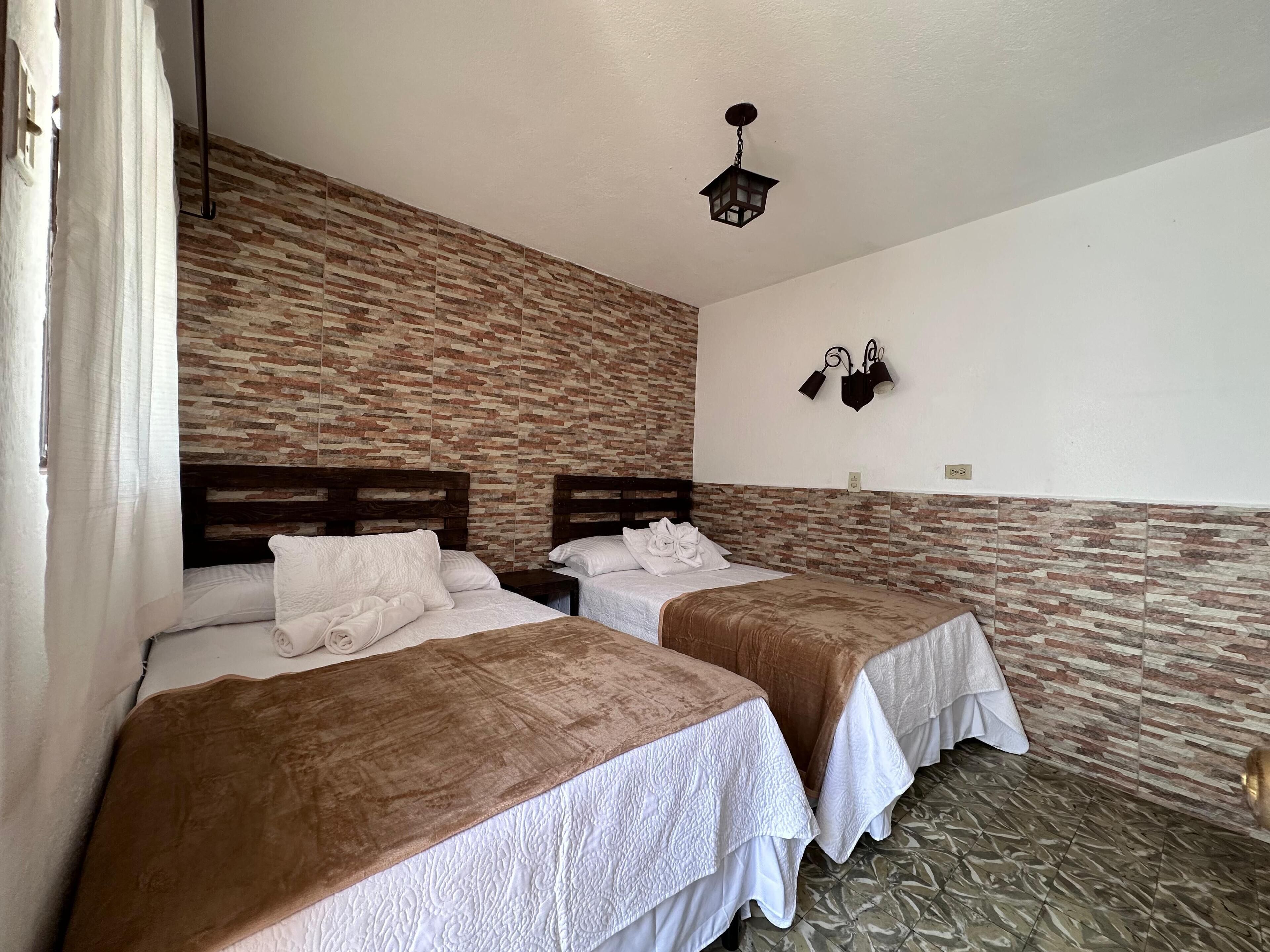 Une chambre avec deux lits, un mur décoré d’un motif en briques, un luminaire suspendu et une petite étagère fixée au mur.