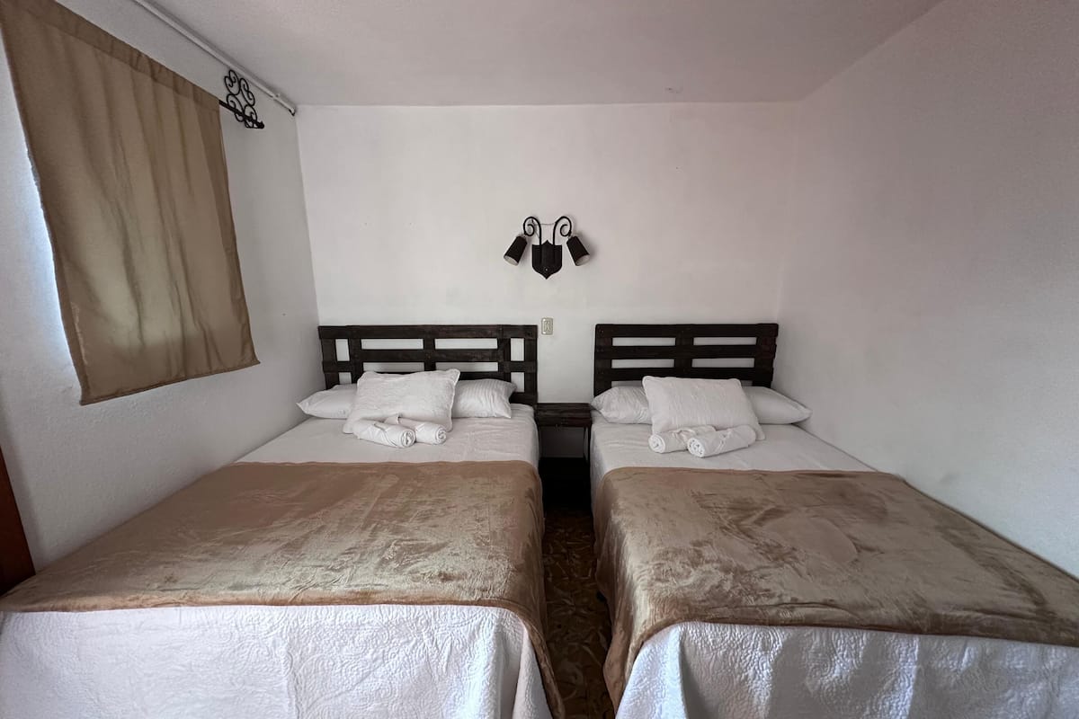 A room with two beds, each with a wooden headboard and white bedding. There is a window with beige curtains, and a wall-mounted light fixture above the beds.