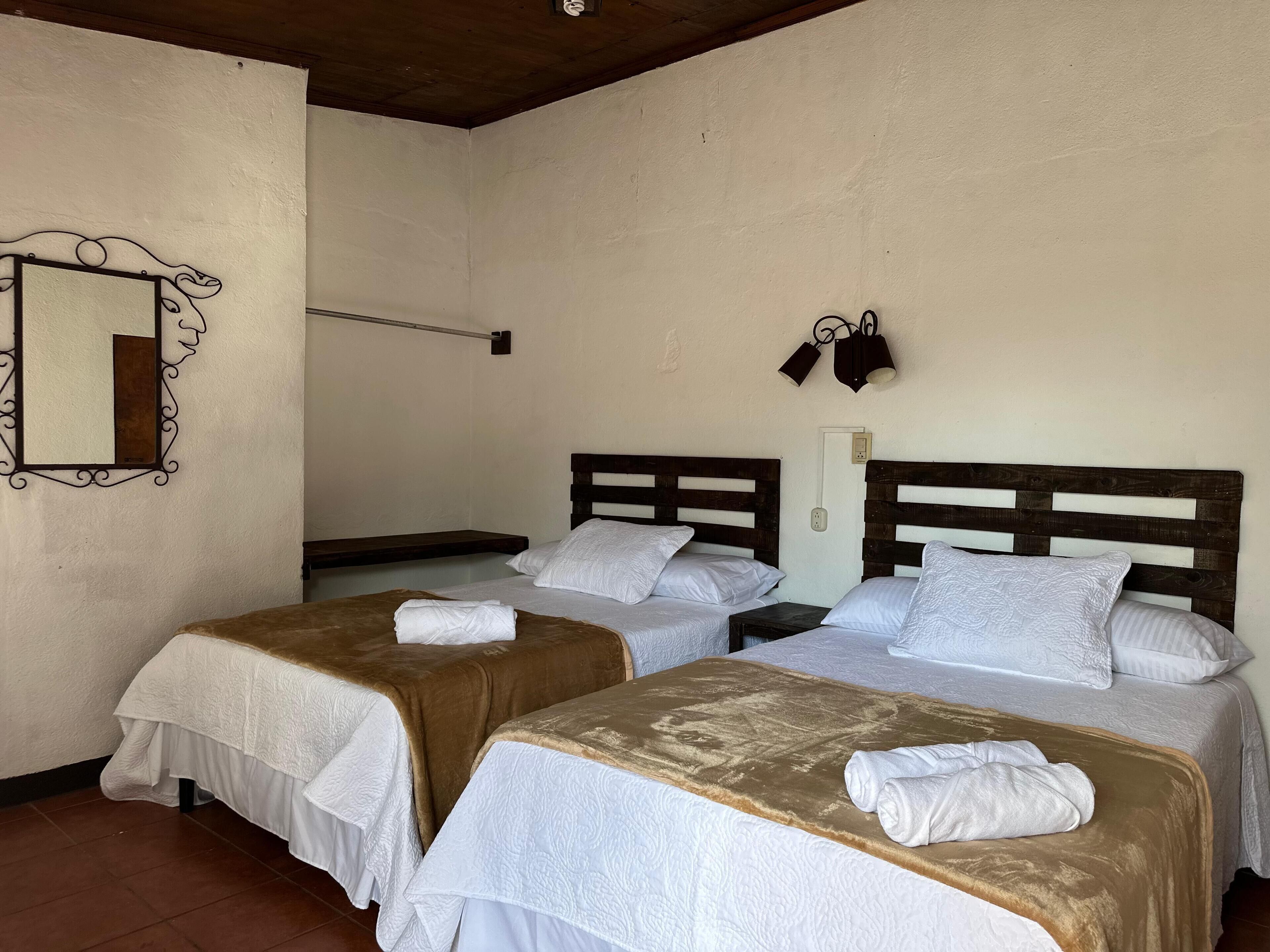Habitación con dos camas, cada una con cabecero de madera y ropa de cama blanca. Un espejo con marco decorativo se encuentra en la pared izquierda, y una lámpara de pared ilumina las camas.