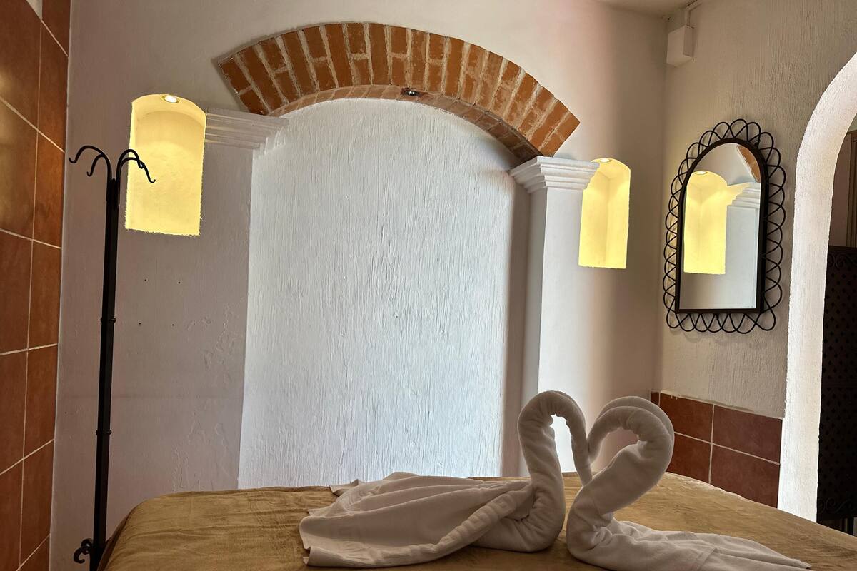 A hotel room with a bed, a brick archway, a wall-mounted mirror, and a floor lamp.