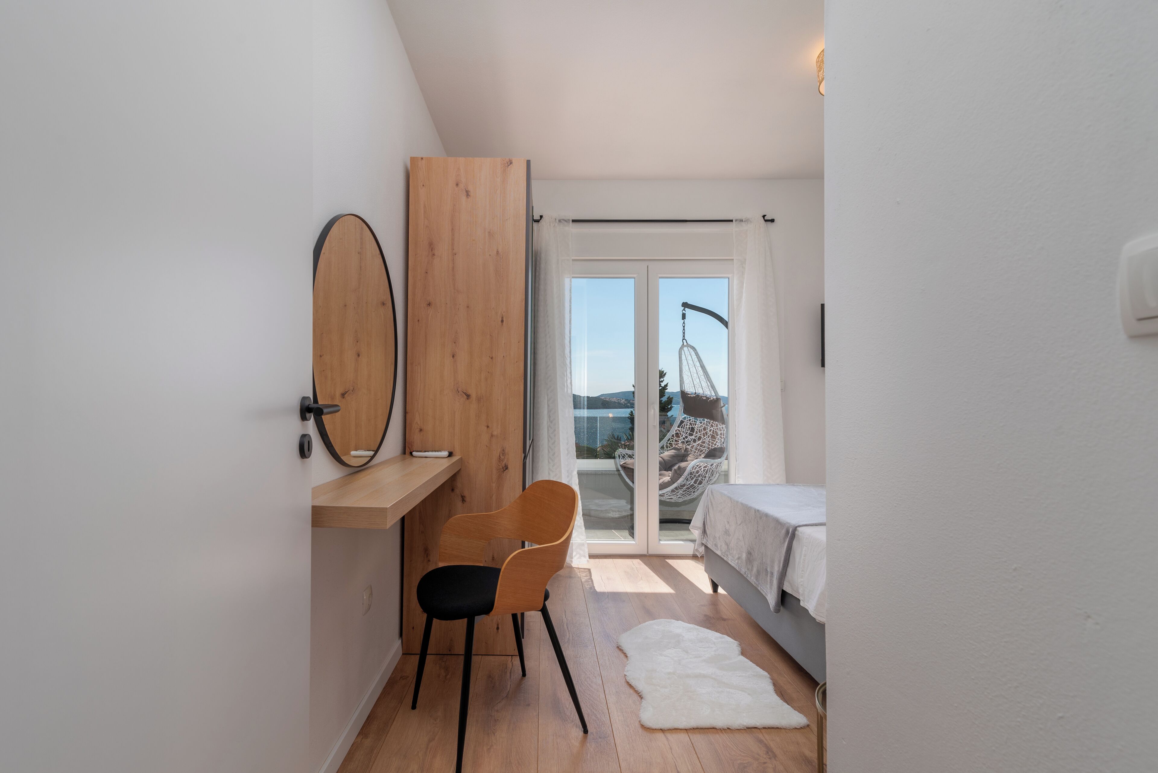 Une petite chambre à coucher moderne, dotée d’un bureau en bois, d’une chaise, d’un lit et offrant une vue sur l’extérieur grâce à une porte-fenêtre coulissante.