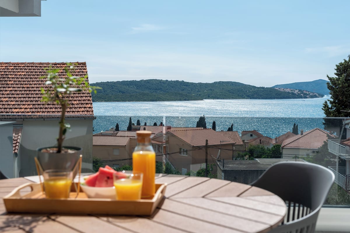 A table with a tray of orange juice and a watermelon, overlooking a lake with houses in the background.