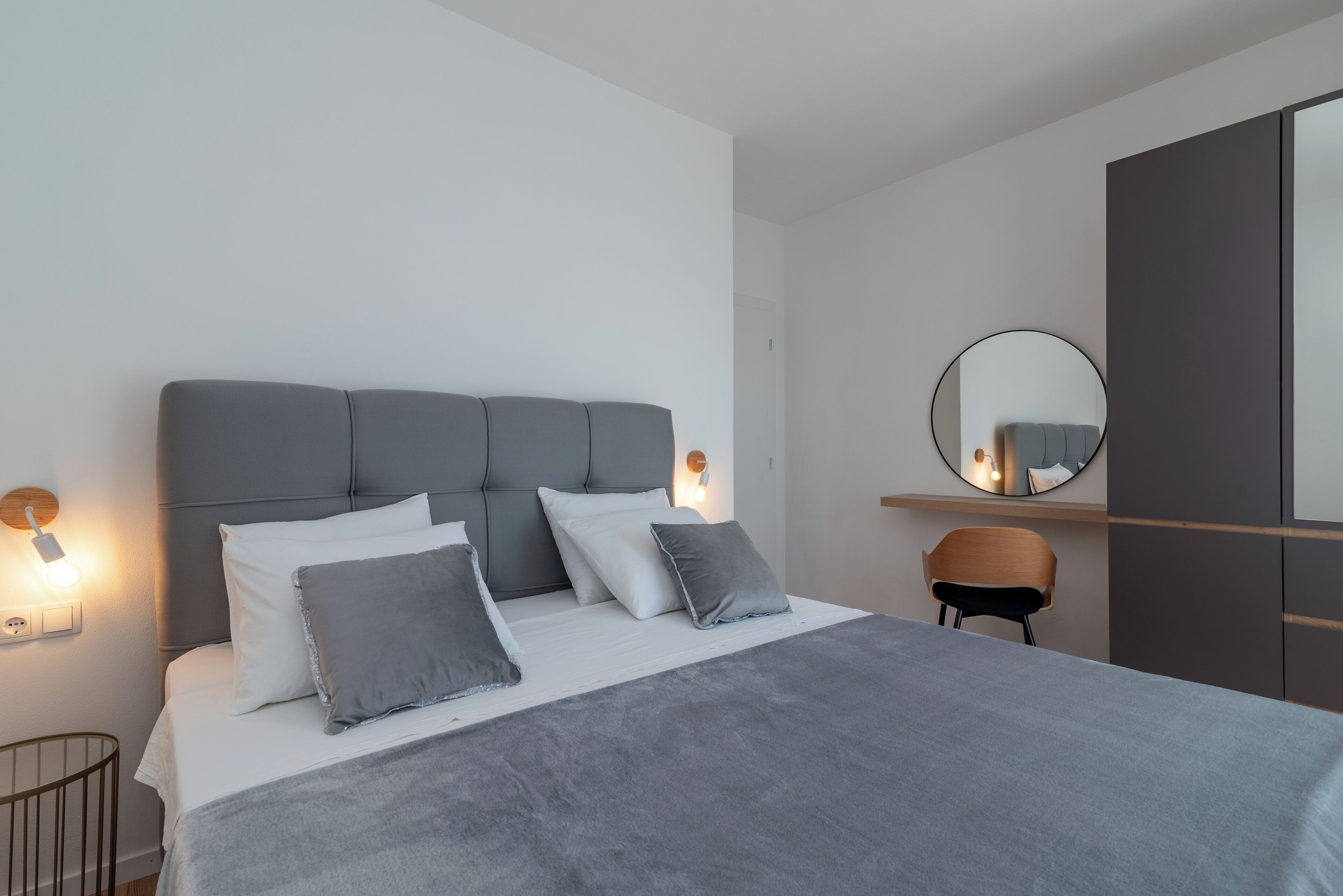 Un dormitorio moderno con un cabecero tapizado en gris, ropa de cama blanca, un espejo redondo y una silla de madera.