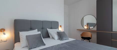 Un dormitorio moderno con un cabecero tapizado en gris, ropa de cama blanca, un espejo redondo y una silla de madera.