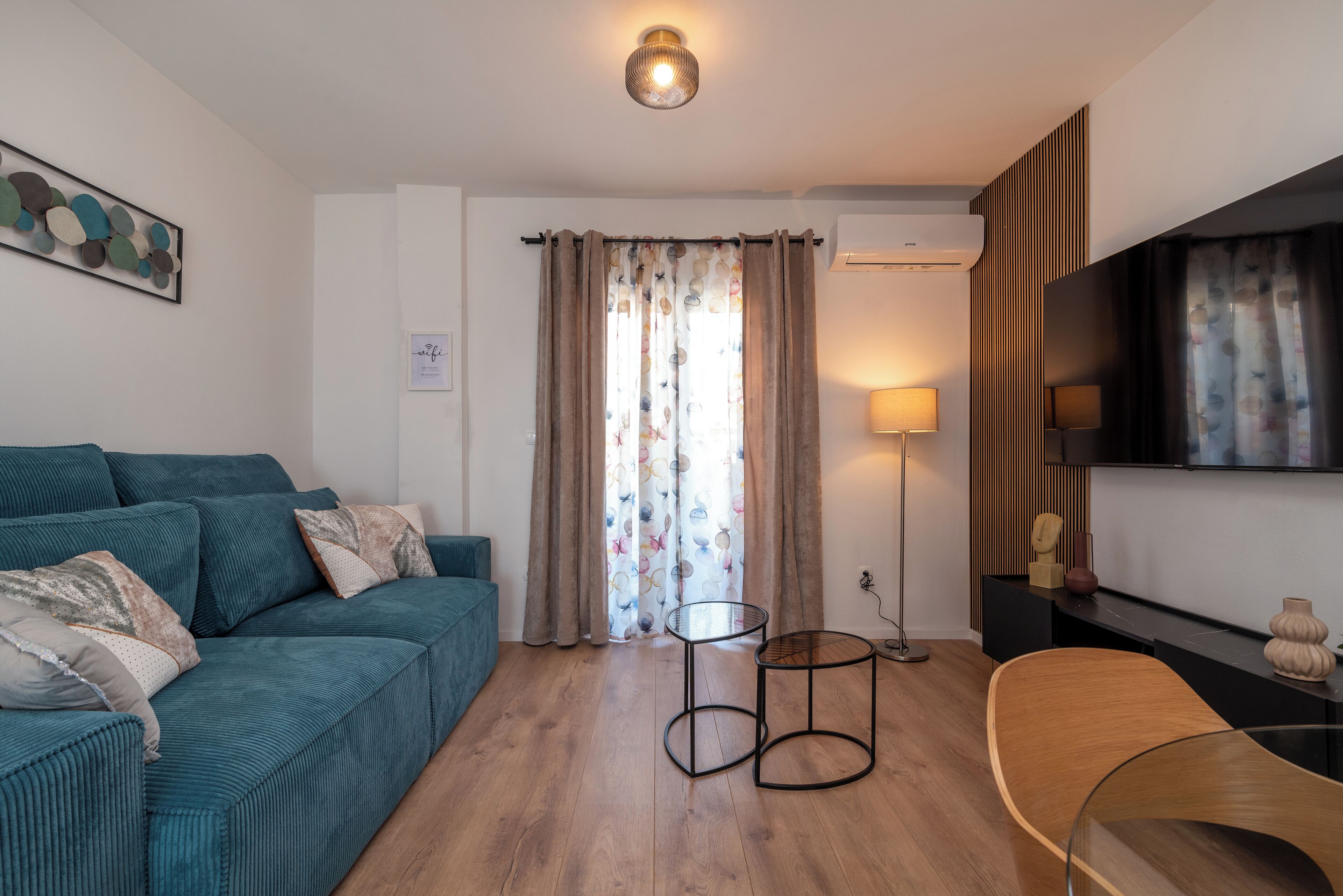 Ein modernes Wohnzimmer mit einem türkisblauen Sofa, einem runden Couchtisch und einem an der Wand befestigten Fernseher.