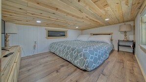 Une chambre avec un plafond en bois, un lit avec une literie à motifs, une table de chevet avec une lampe et une commode.