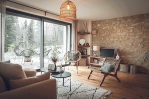 A cozy living room with a stone wall, large windows, wicker furniture, and a wooden floor. - Gite plain pied dans ancienne ferme en pierre - bain nordique et sauna (Prémanon)