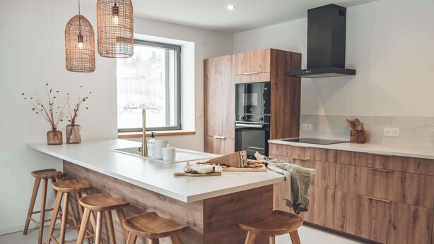 A modern kitchen with a large island, wooden stools, pendant lights, and a window providing natural light. - Gîte dans ferme en pierre, bain nordique et sauna (Prémanon)