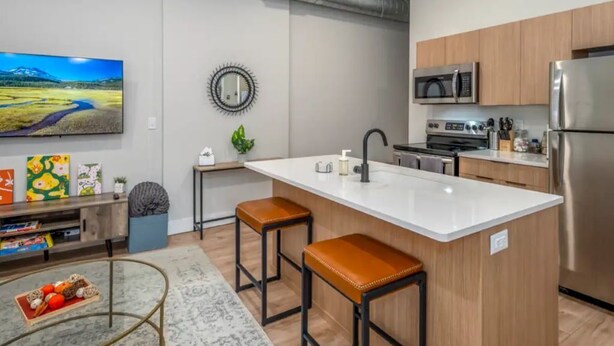 A modern kitchen with a central island, wooden cabinets, stainless steel appliances, and a flat-screen TV mounted on the wall. - OTR, 3 Lux Queens, Two 55in TVs,  by TQL, Findlay Mkt, FREE Light Rail, Sleeps 7 (Cincinnati)