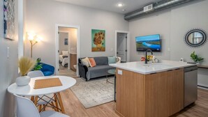 A modern kitchen with a central island, a dining area with a white table and chairs, and a living area with a sofa and a TV. - OTR, 3 Lux Queens, Two 55in TVs,  by TQL, Findlay Mkt, FREE Light Rail, Sleeps 7 (Cincinnati)