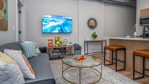 A modern living room with a grey sofa, a glass coffee table, and a TV mounted on the wall. - OTR, 3 Lux Queens, Two 55in TVs,  by TQL, Findlay Mkt, FREE Light Rail, Sleeps 7 (Cincinnati)