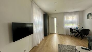 Una sala de estar moderna con un televisor de pantalla plana, un comedor con mesa y sillas, y un ventanal con cortinas de gasa.