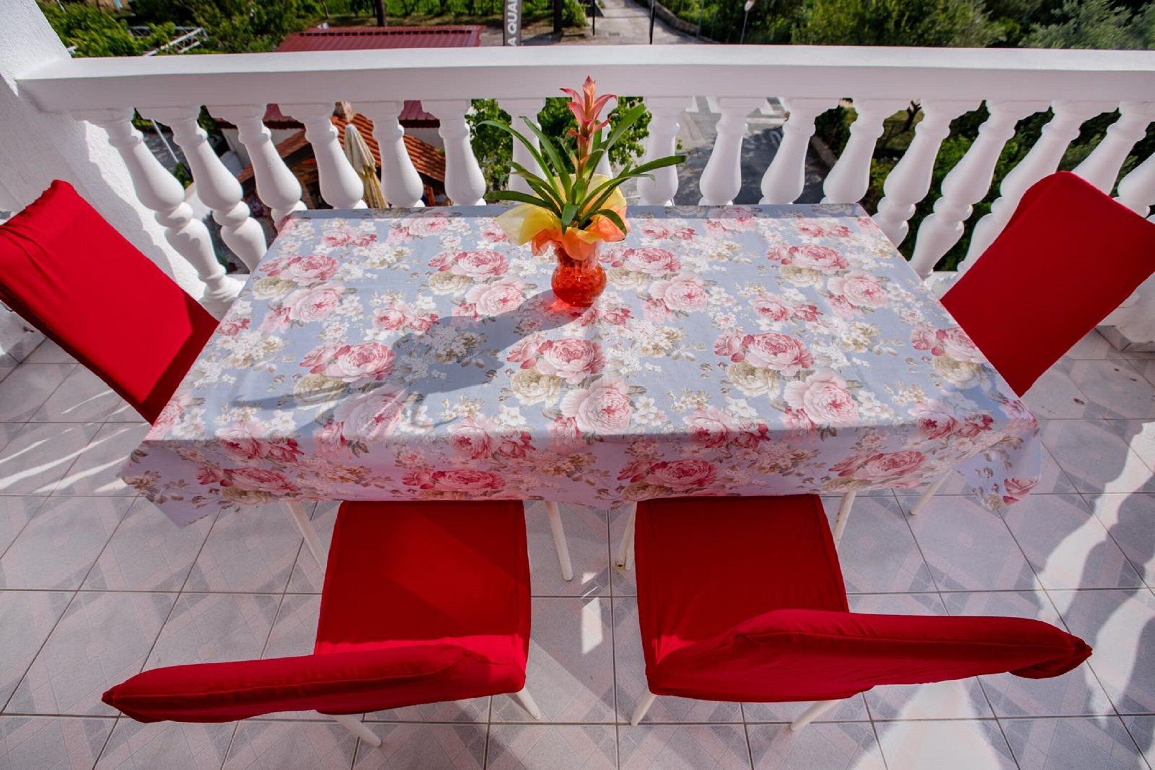 Une table carrée recouverte d’une nappe à motifs floraux, avec des coussins rouges et un vase rempli de fleurs.