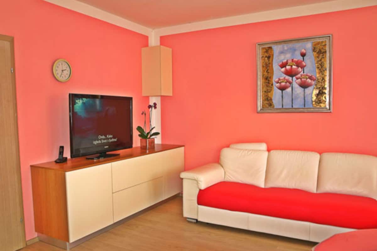A room with a pink wall, a television on a wooden cabinet, a white sofa with a red cushion, and a framed picture above the sofa.