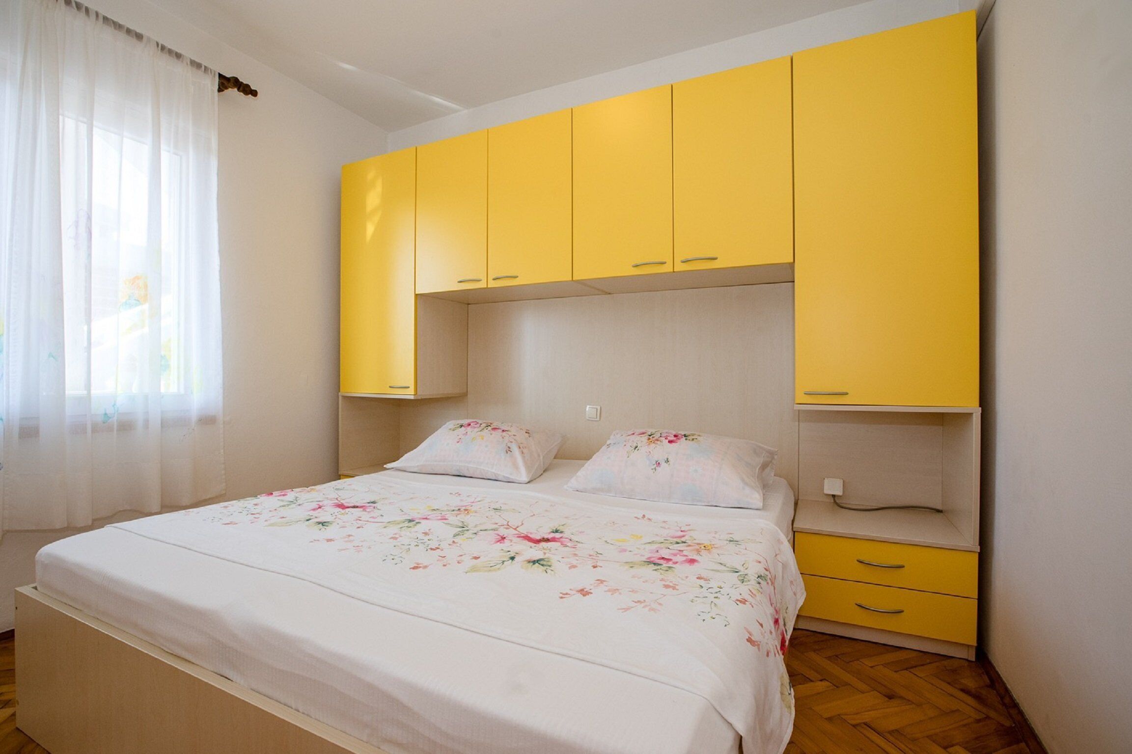 A bedroom with a bed, floral bedding, yellow cabinets, and a window with sheer curtains.