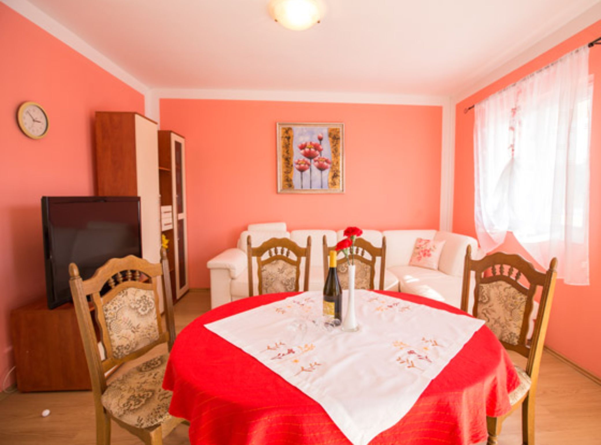 A dining area with a table set for four, surrounded by wooden chairs with floral upholstery. A television is placed on a cabinet to the left.