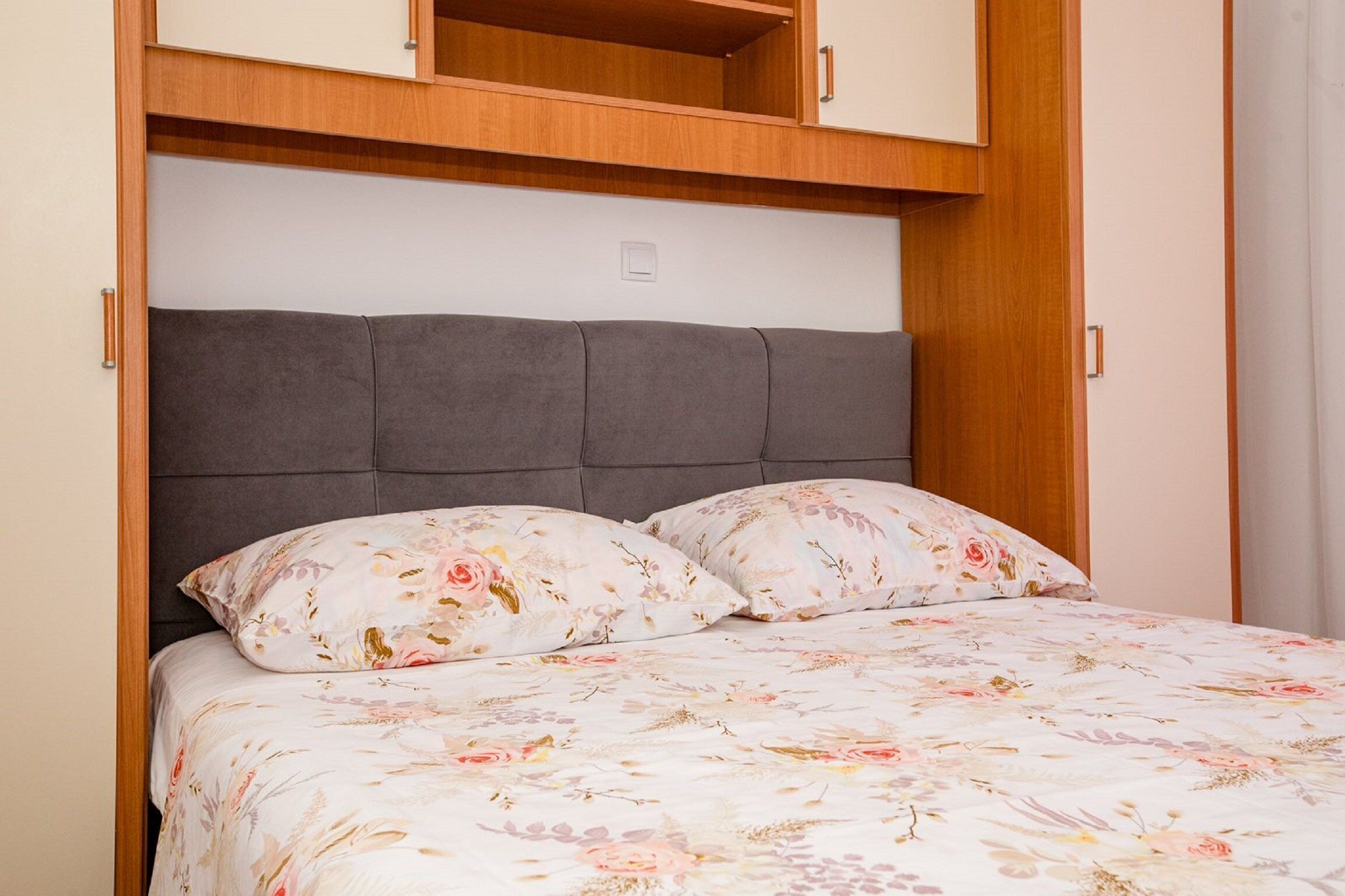 Una camera da letto con un letto, biancheria da letto floreale, una testiera imbottita grigia e mobili contenitori in legno sopra.