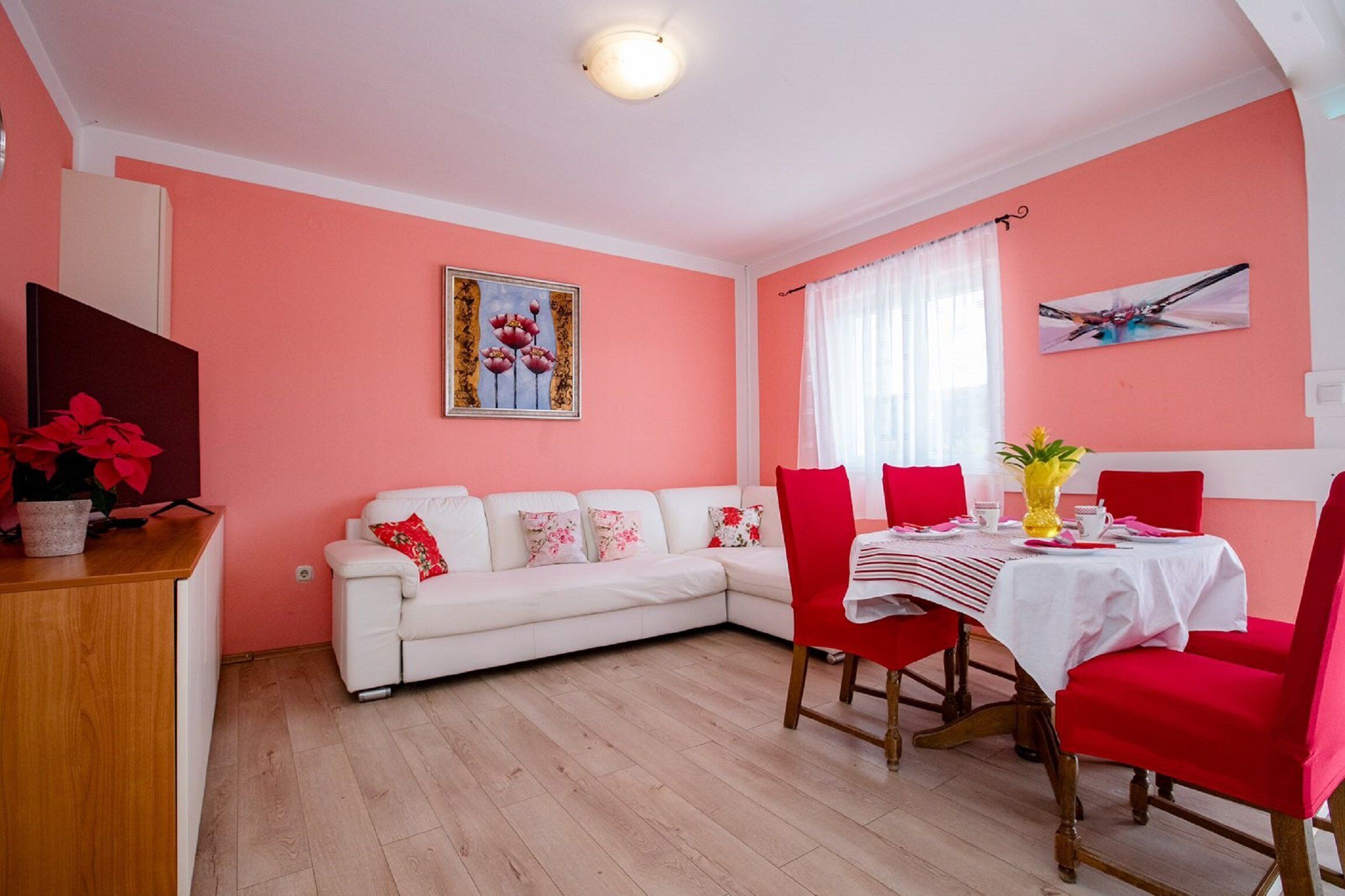 Un salon avec un canapé blanc, des fauteuils rouges, une table à manger recouverte d’une nappe blanche et un vase rempli de fleurs.