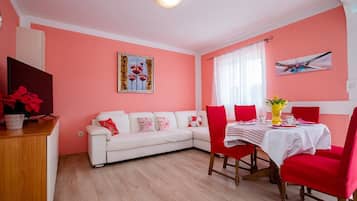 Una sala de estar con un sofá blanco, sillas rojas, una mesa de comedor con mantel blanco y un jarrón con flores.