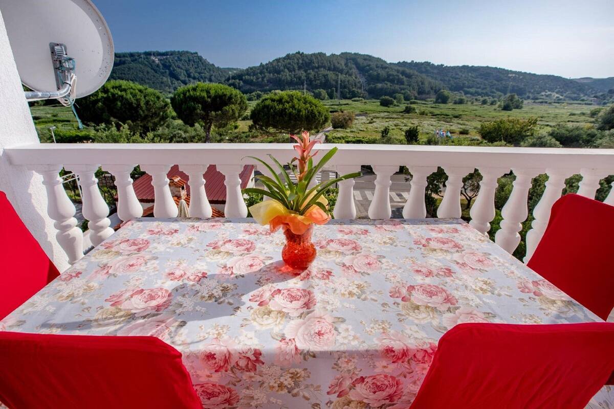 A balcony with a table set for two, a vase with flowers, and a scenic view of a green landscape.