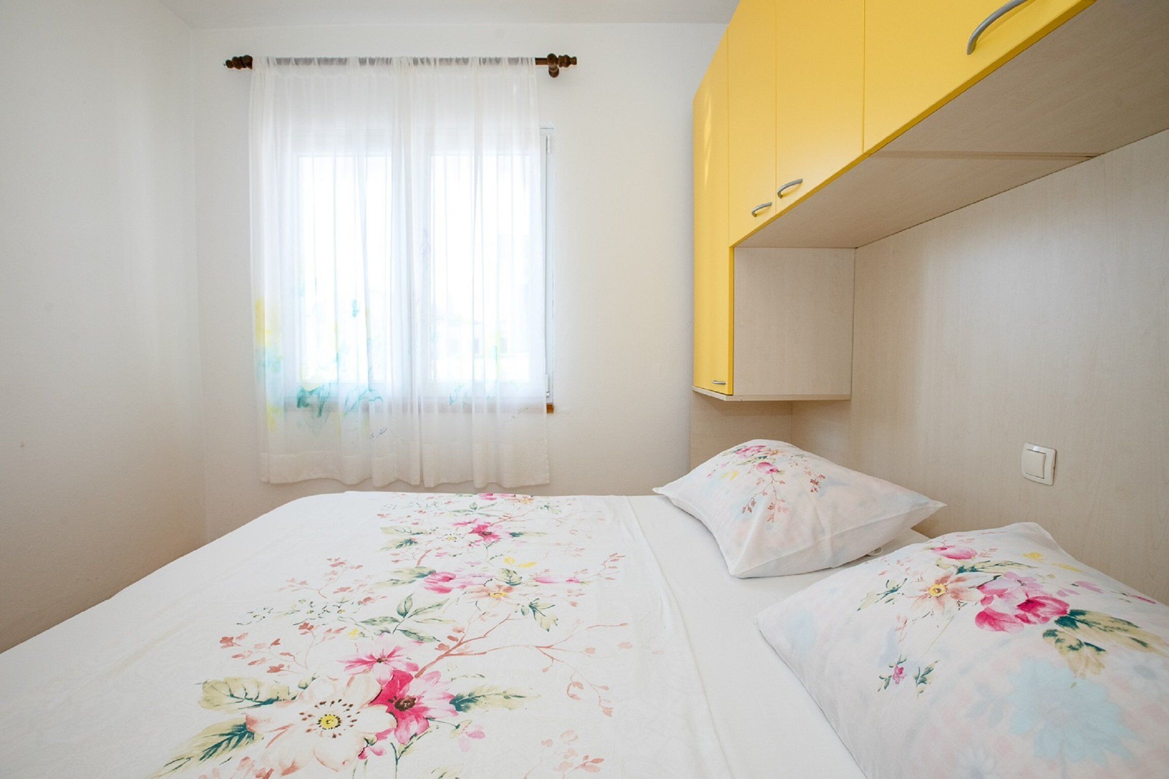 Quarto com cama, roupa de cama florida, janela com cortinas leves e guarda-roupa amarelo.