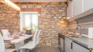 A compact kitchen with stone walls, a dining area with white chairs, and a window with natural light.