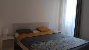 Un lit bien fait, recouvert d’une couvre-lit à motifs, une lampe de chevet et une fenêtre ornée de rideaux légers.