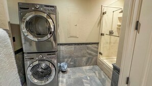Un espace buanderie comprenant deux machines à laver et deux sèche-linge superposés, une douche et un sol carrelé.