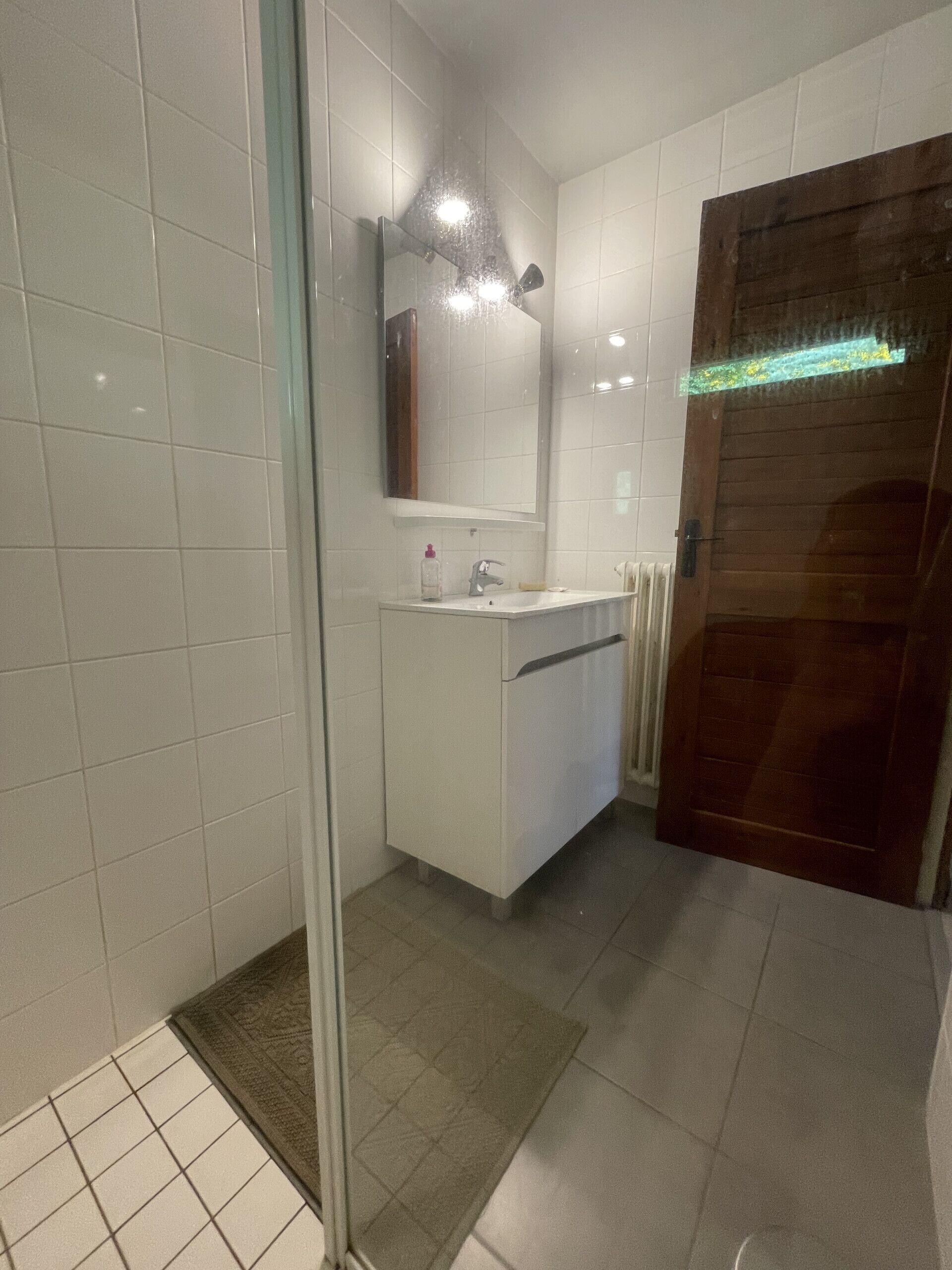 A bathroom with white tiles, a glass shower enclosure, a wooden door, and a white sink with a mirror above it.