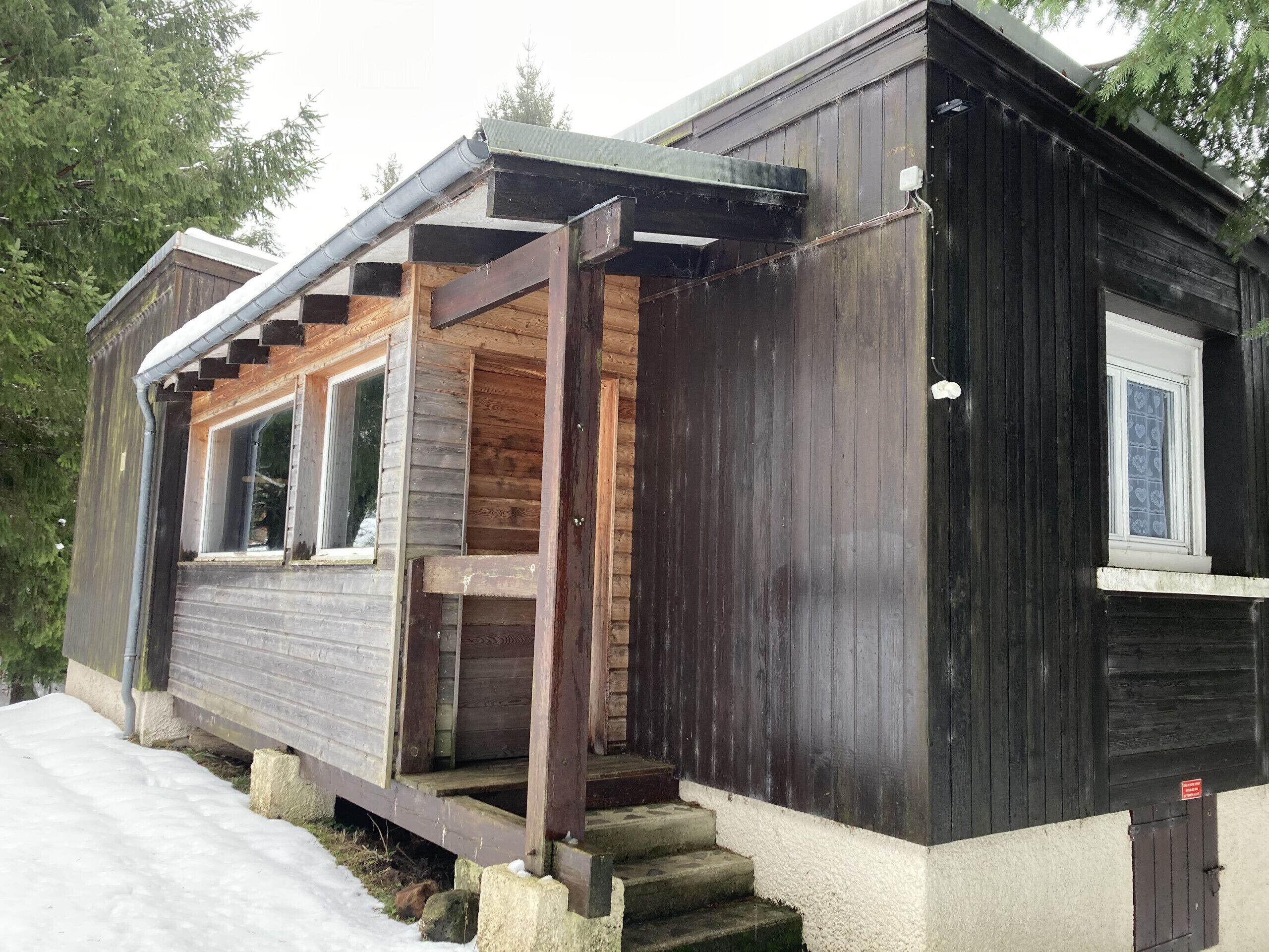 Une cabane avec une façade en bois, une véranda et une fenêtre ornée d’un rideau à motifs de cœur.