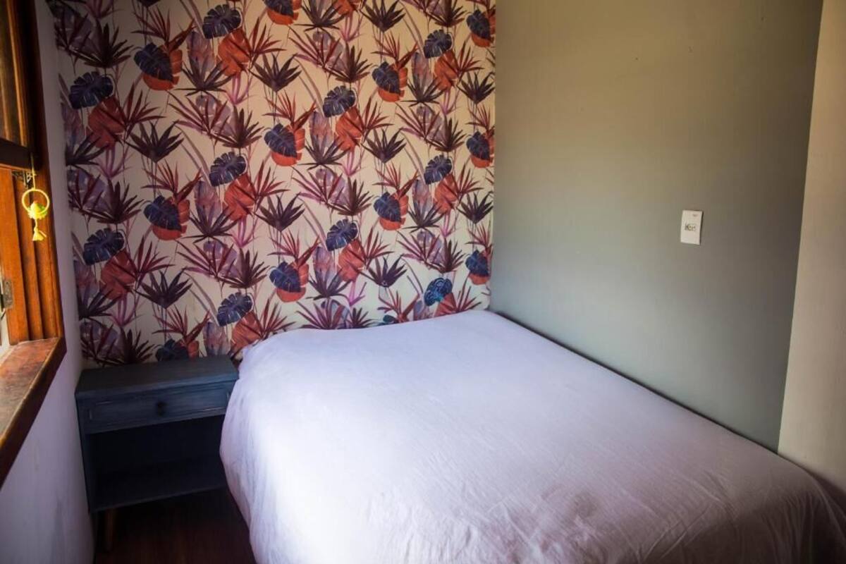 A small, single-bed room with a floral-patterned wallpaper, a wooden nightstand, and a wall socket.