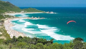 A coastal landscape with a beach, rocky outcrops, and a parasail in the distance. - Island Vibe Nomads - Adventure Hostels (Florianópolis)