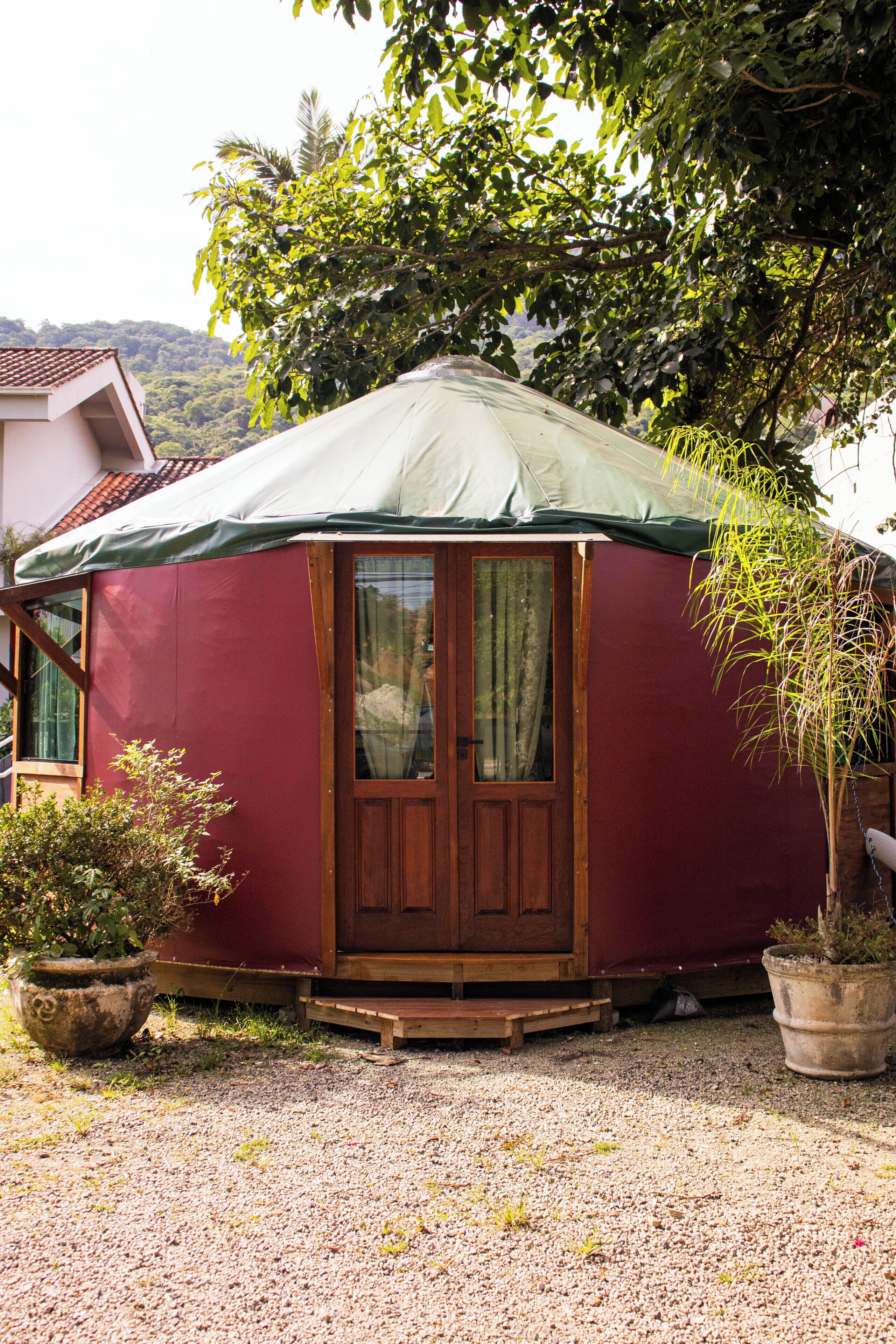 Una yurta con techo verde, puerta de madera y ventanas. Está rodeada de grava y hay plantas en macetas cerca.