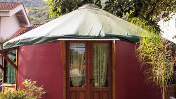 Una yurta con techo verde, puerta de madera y ventanas. Está rodeada de grava y hay plantas en macetas cerca.