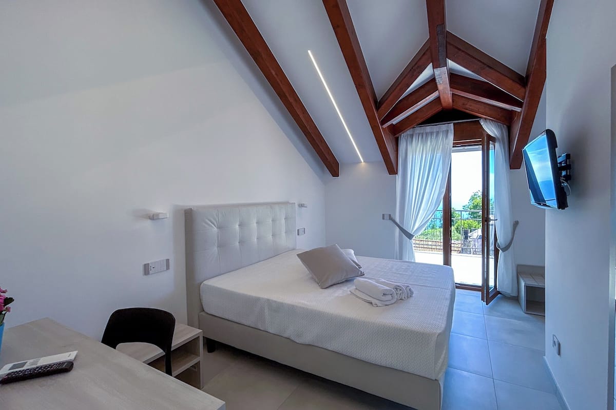 A bedroom with a bed, a desk, a chair, a TV, and a balcony with curtains.