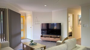 Una sala moderna con una televisión de pantalla plana sobre un soporte de madera, un sofá, una mesa de centro con una planta, y una puerta con un cartel de 'Welcome'.
