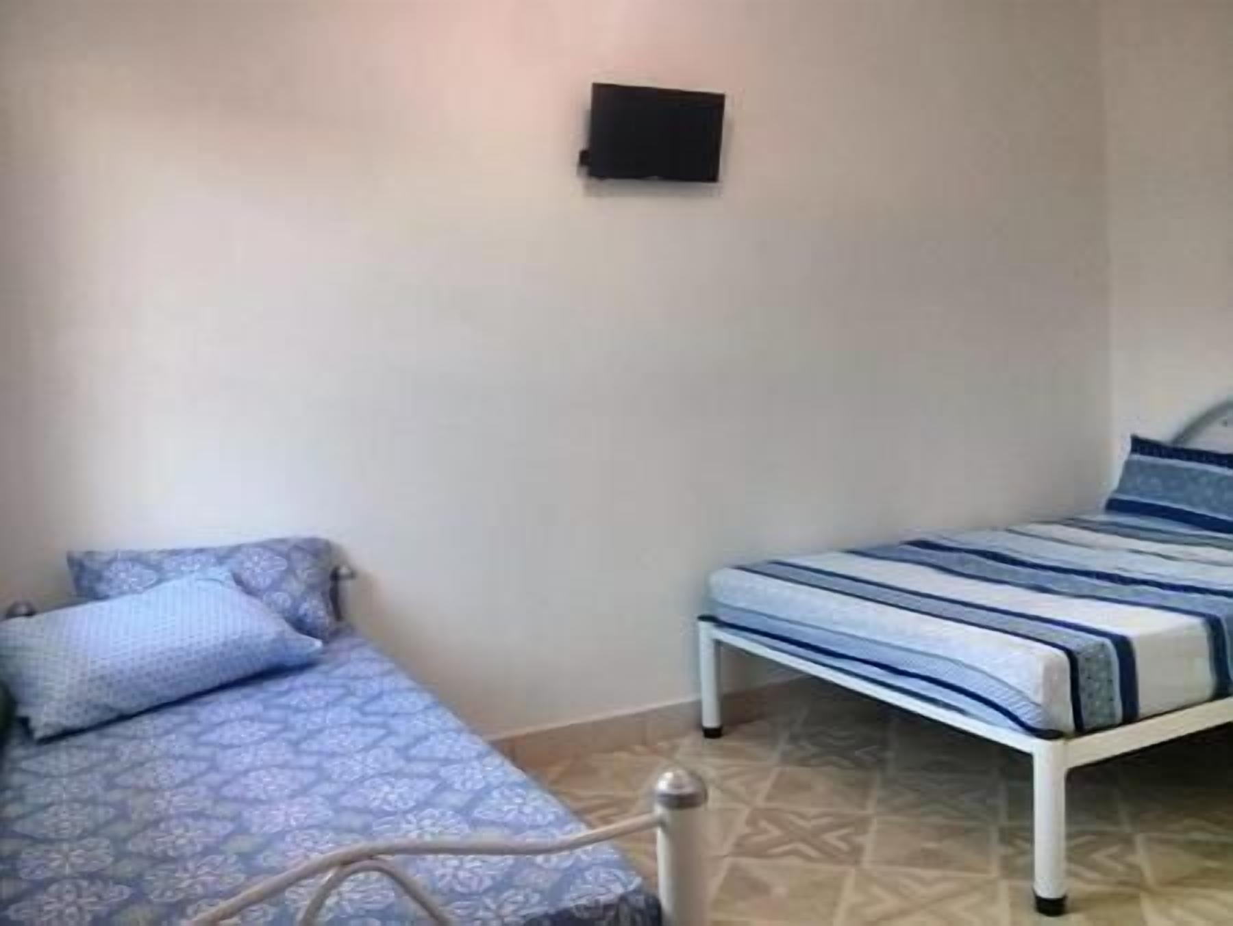 A small, simple room with two beds, one with a patterned blue and white bedspread and the other with a striped blue and white bedspread. There is a small black object on the wall above the beds.