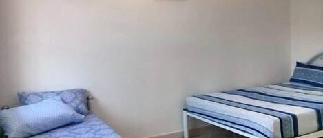 A small, simple room with two beds, one with a patterned blue and white bedspread and the other with a striped blue and white bedspread. There is a small black object on the wall above the beds.
