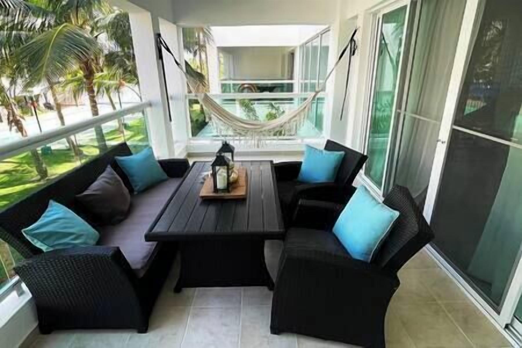 A covered outdoor seating area with a hammock, black cushioned chairs, and a central table with a lantern.